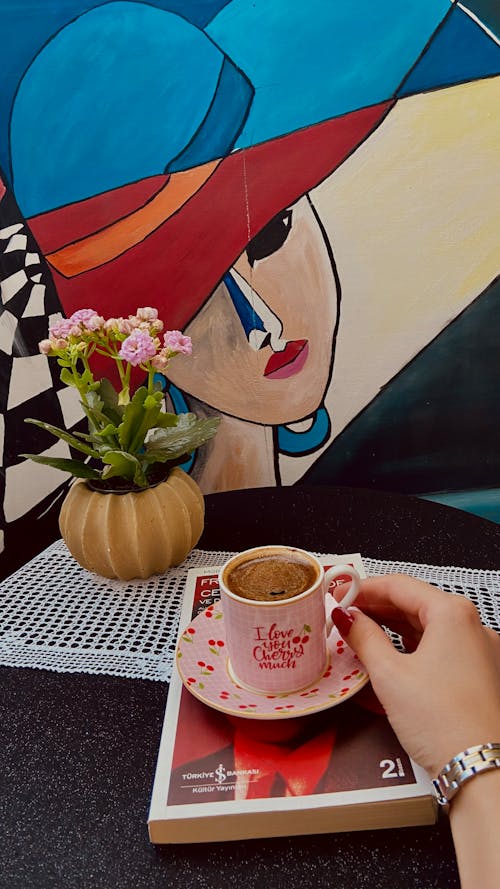 Woman Hand on Coffee Cup on Book near Painting on Wall