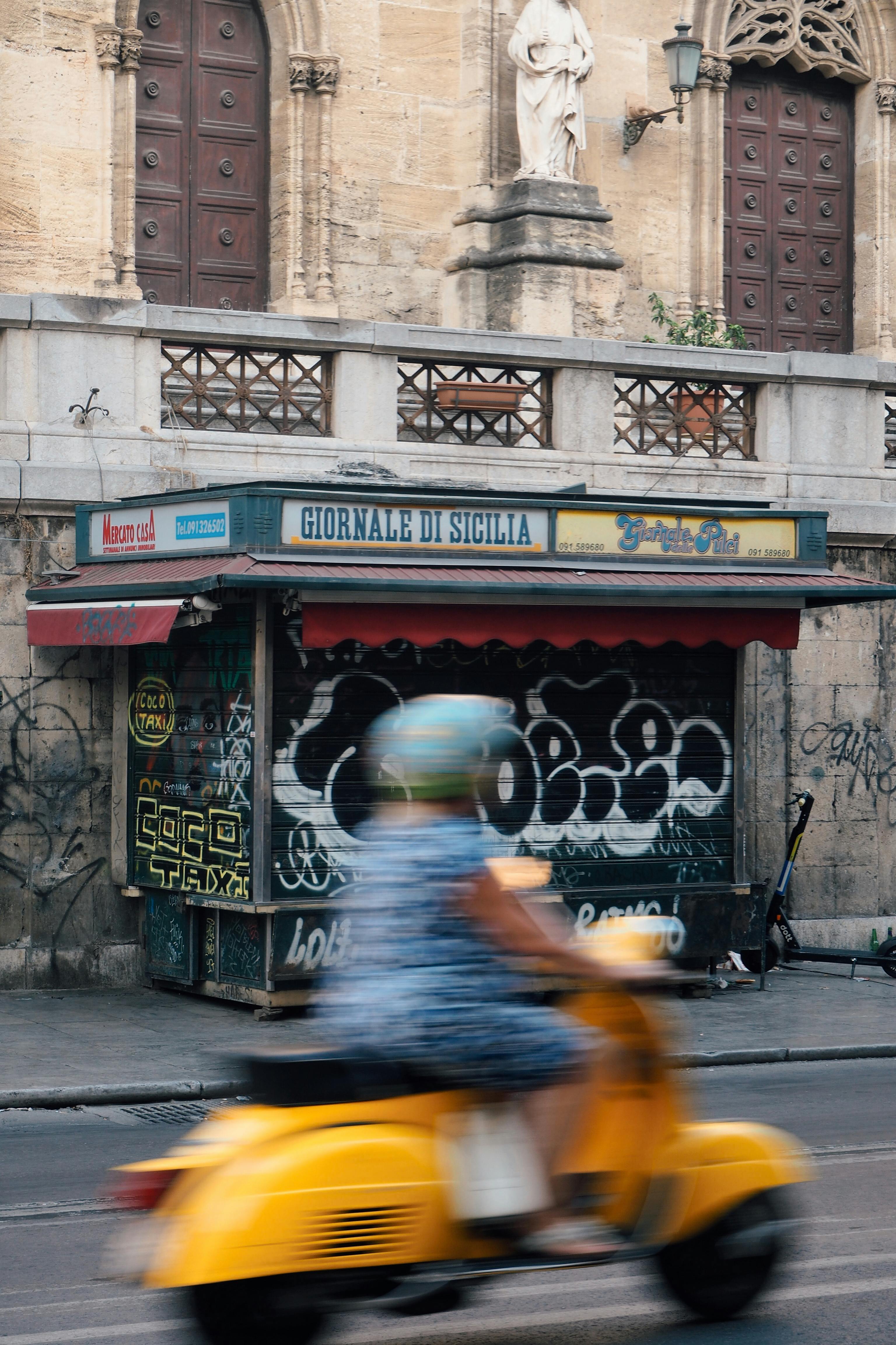 A yellow scooter blurs past a graffiti-covered newsstand in Sicily's urban streets.