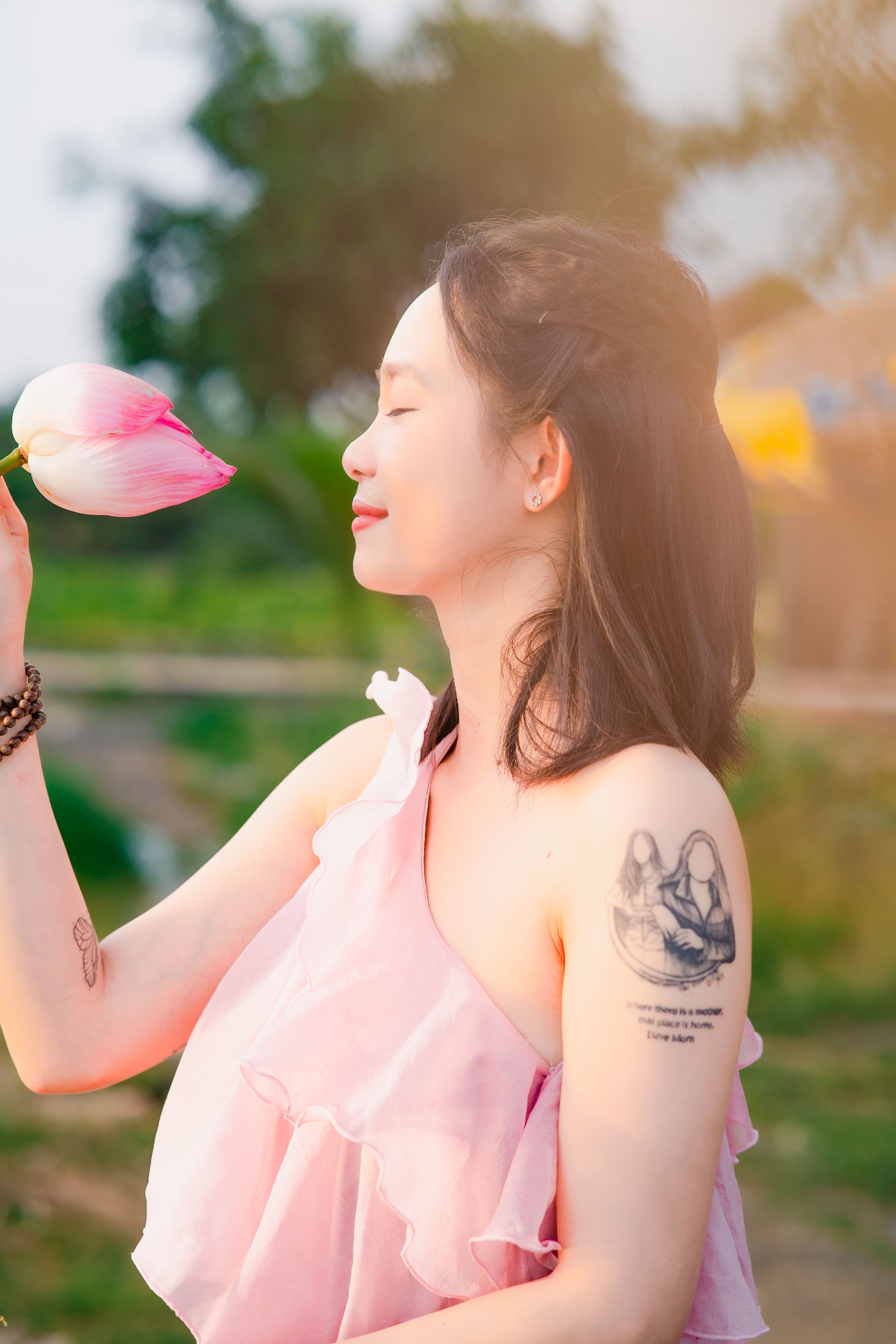 A peaceful woman with tattoos embraces nature while holding a pink lotus flower outdoors.