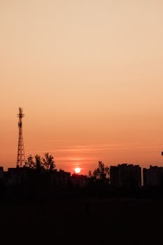 Silhouette of urban buildings and a tower against a warm sunset sky.