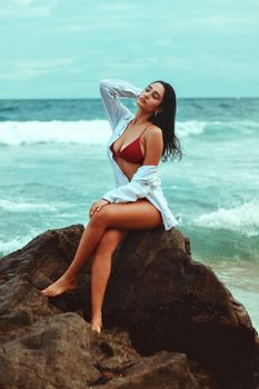 Confident woman in red bikini with white shirt posing on seaside rocks with ocean waves in the background.