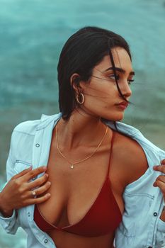 Stylish portrait of a woman in a red bikini and white shirt by the sea.
