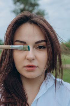 Close-up portrait of a young woman holding a paintbrush outdoors, showcasing natural beauty and creativity.
