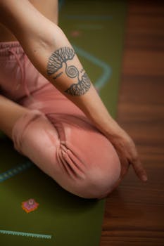 A woman meditating cross-legged with a tree tattoo on her arm. Calming indoor yoga atmosphere.