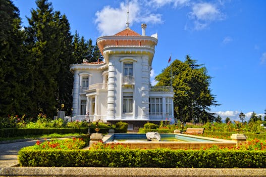 A beautiful white mansion surrounded by lush gardens and a fountain on a sunny day.