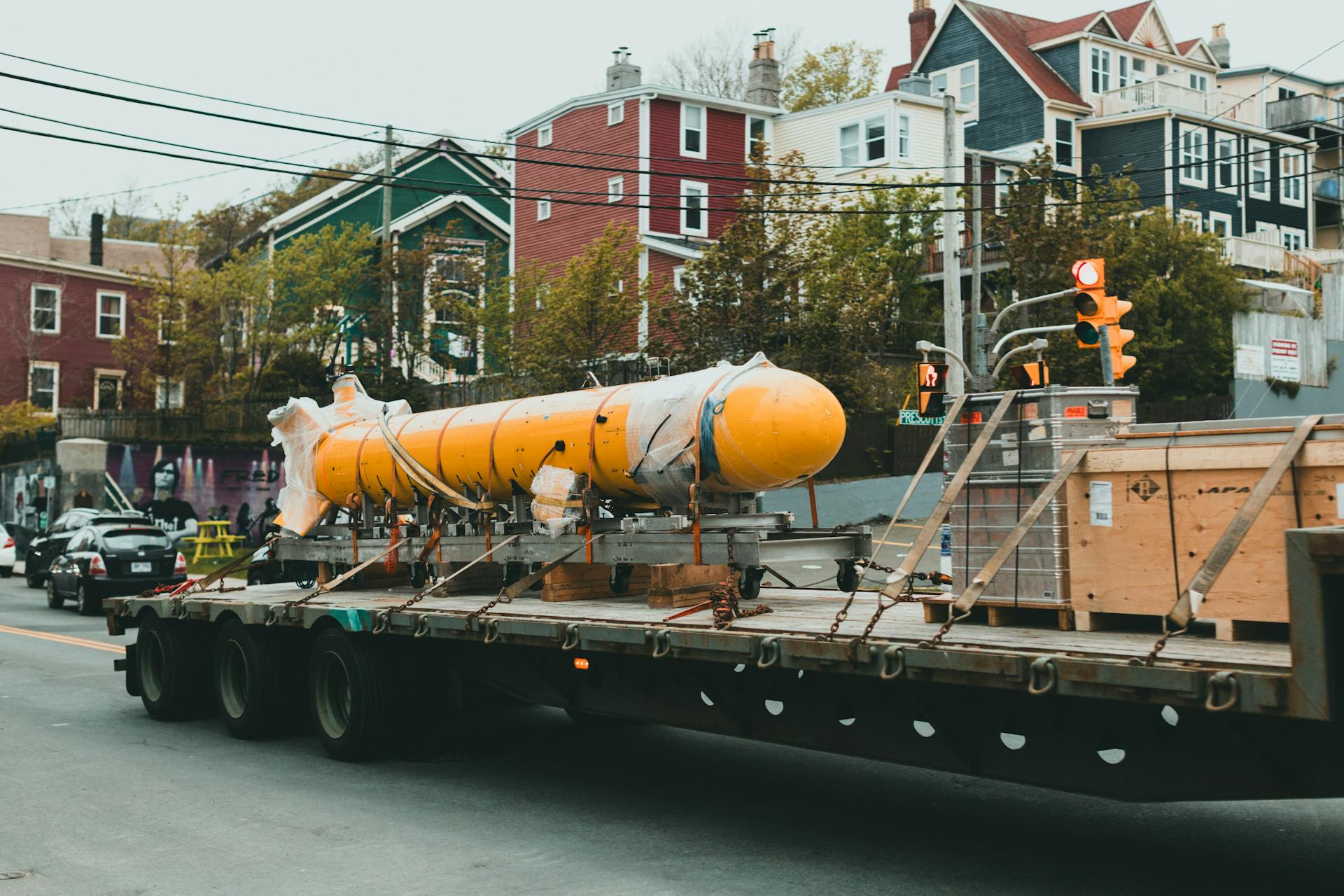 A large yellow rocket is transported on a flatbed truck through a city street, surrounded by buildings and trees.