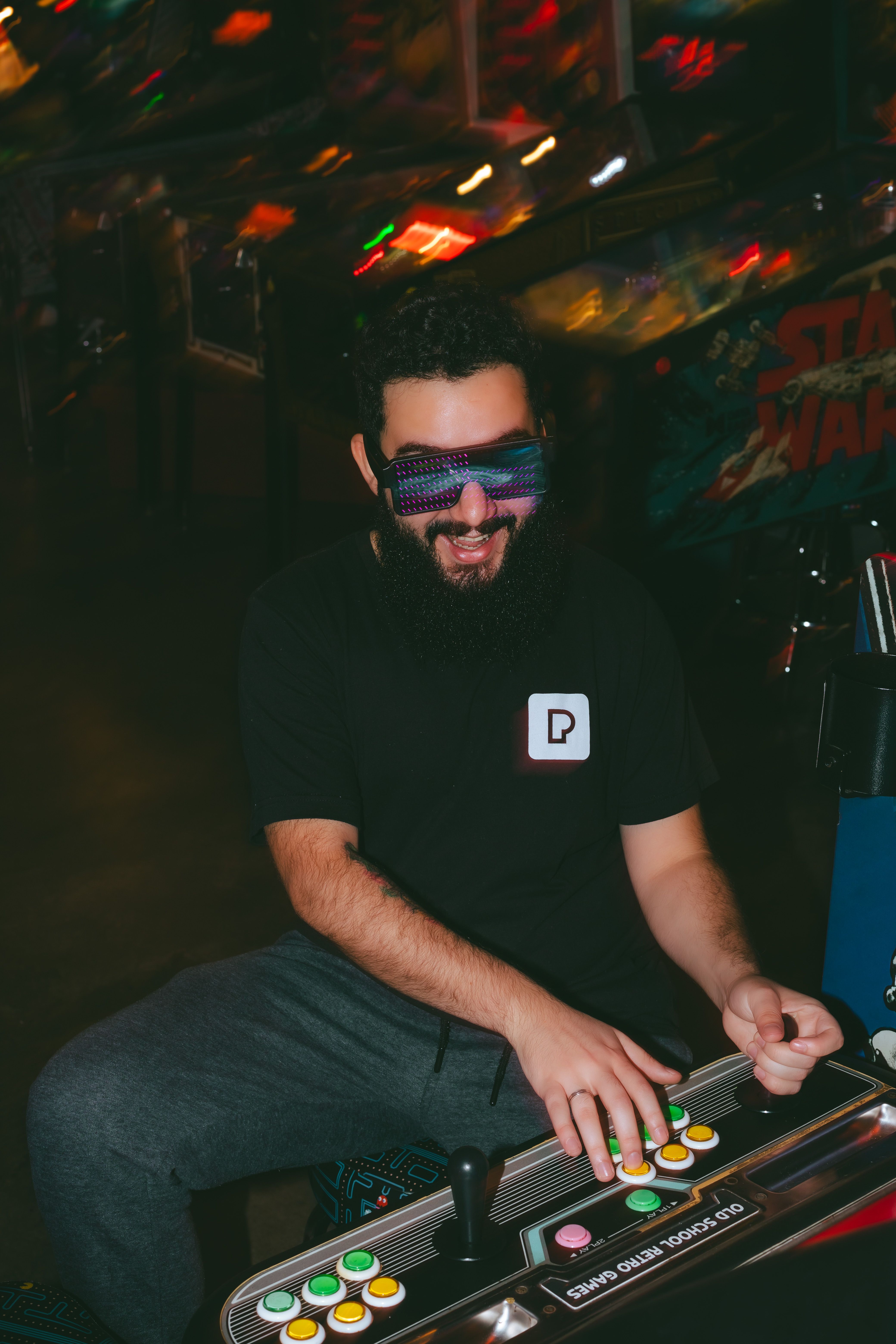 Smiling man with beard in black t-shirt plays colorful arcade machine wearing futuristic sunglasses.