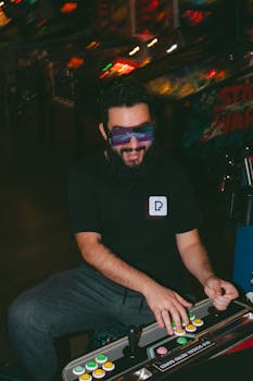 Smiling man with beard in black t-shirt plays colorful arcade machine wearing futuristic sunglasses.