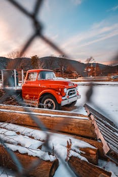 A vintage red truck parked by logs and snow at Lake George, NY, with a scenic mountain backdrop.