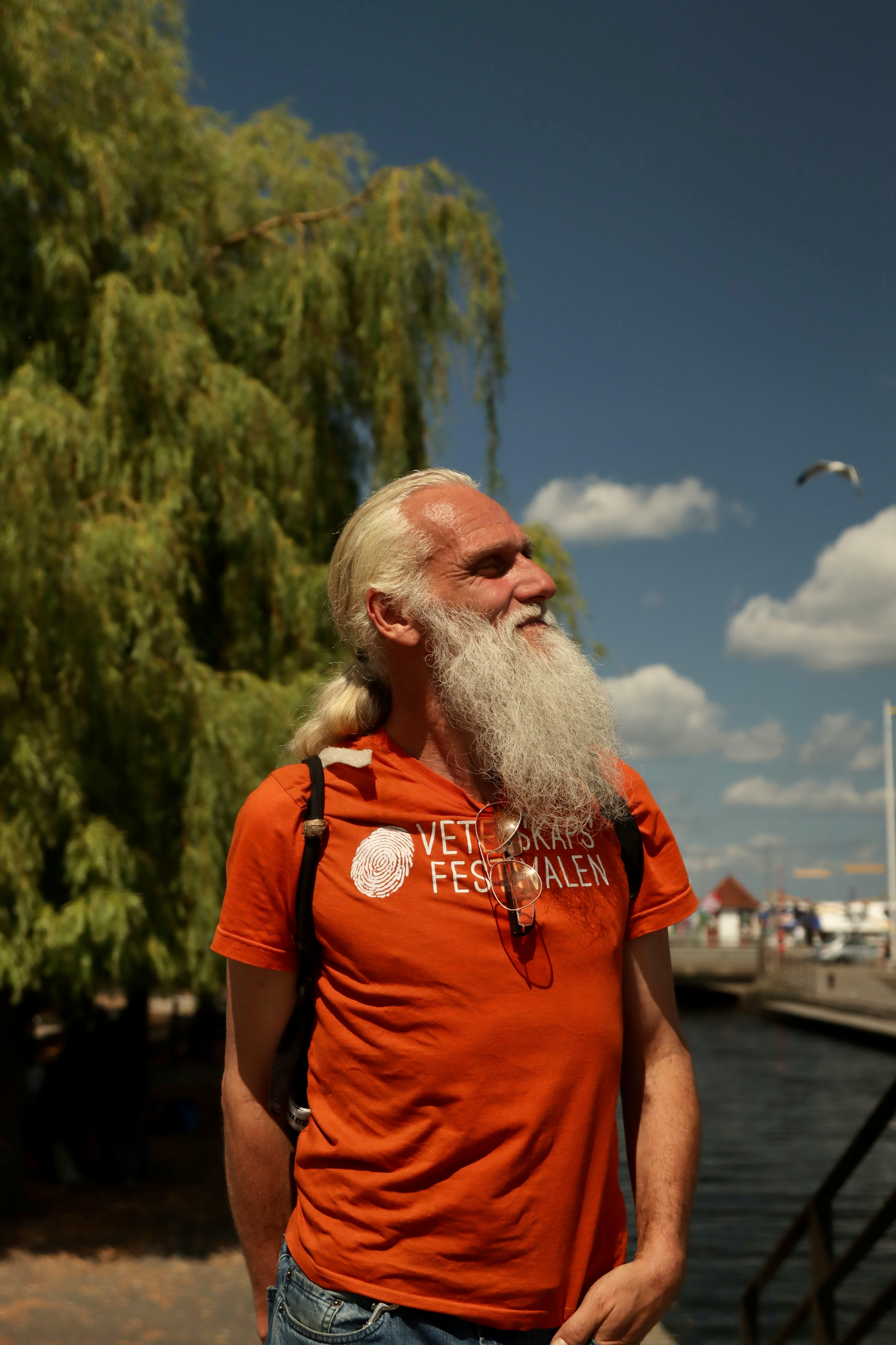 Elderly man with long white beard and orange t-shirt standing by the waterfront in Jönköping, Sweden, on a sunny day.