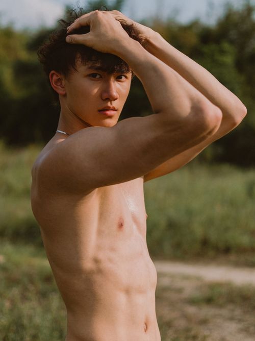 Free Shirtless young man standing in a natural setting with arms raised, showcasing a confident and relaxed pose. Stock Photo