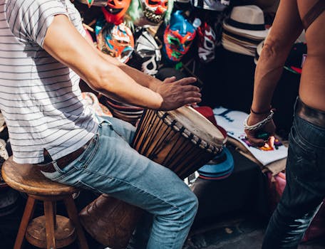 Musician performs on a djembe drum at a colorful outdoor market with vibrant masks.