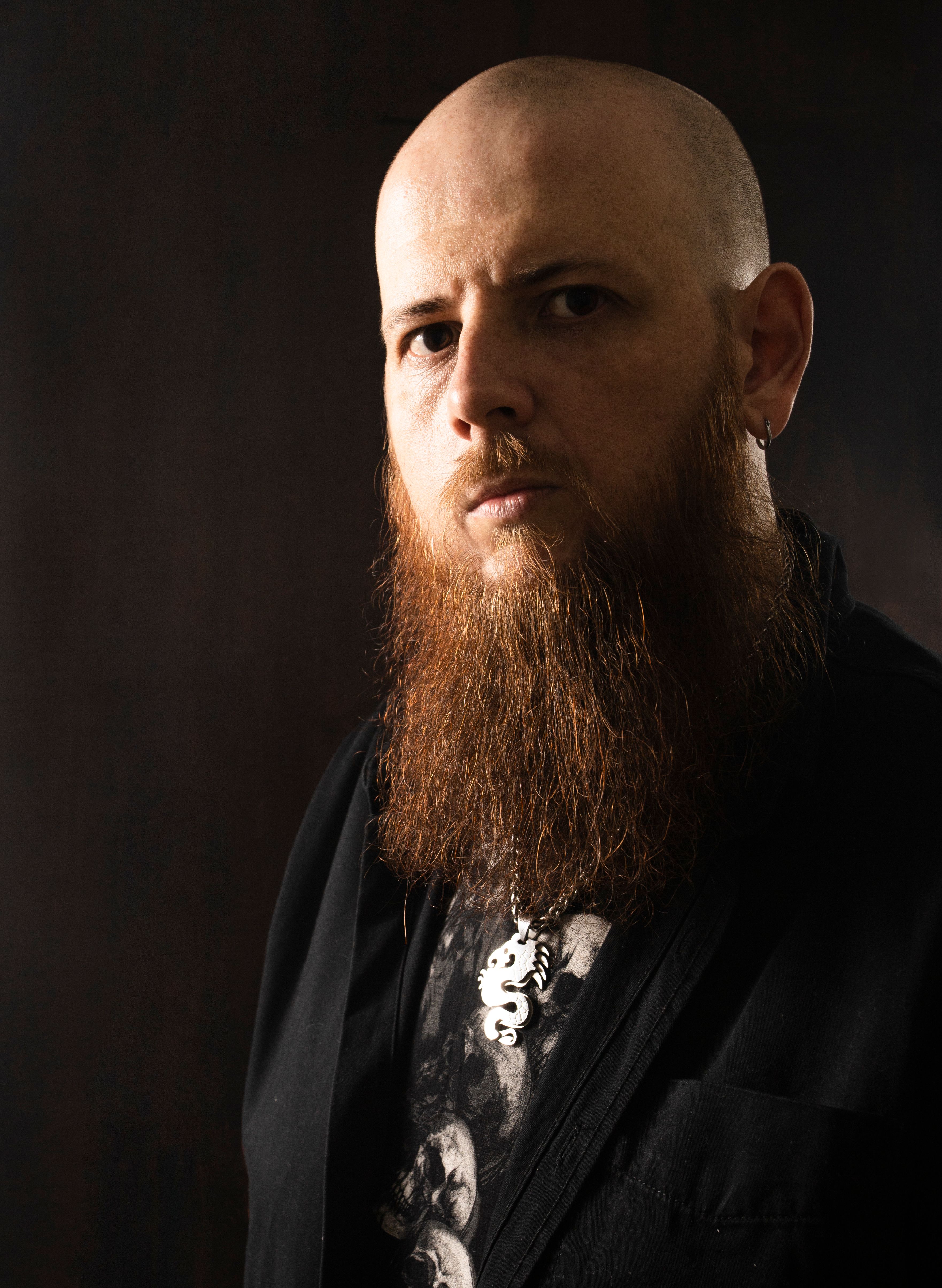 Serious portrait of a bald man with a ginger beard and necklace in studio lighting.
