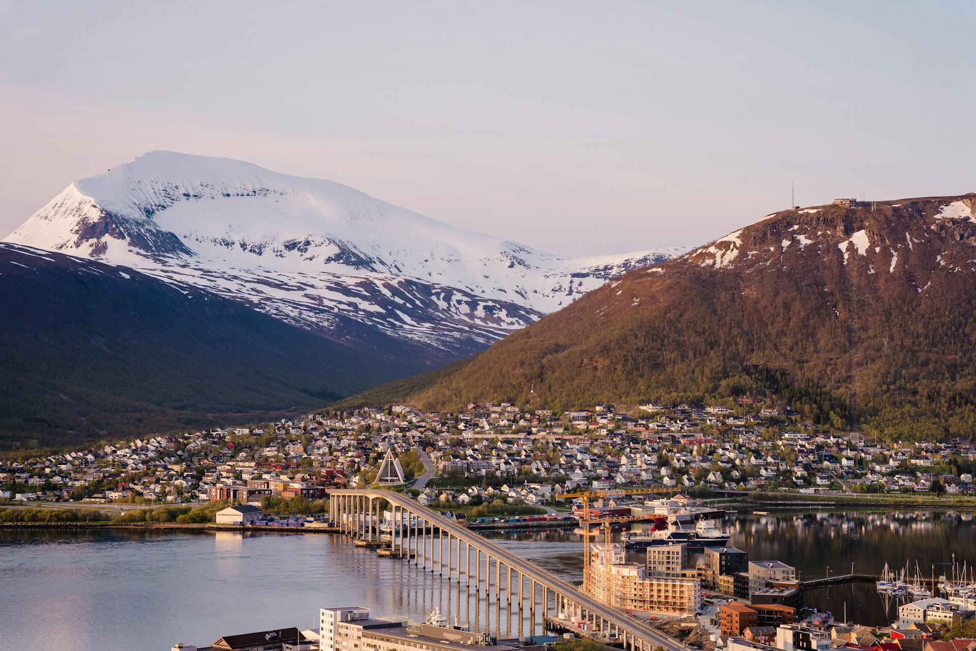 Aerial view of Tromsø bridge, city, and snowcapped mountains during sunset in Norway.