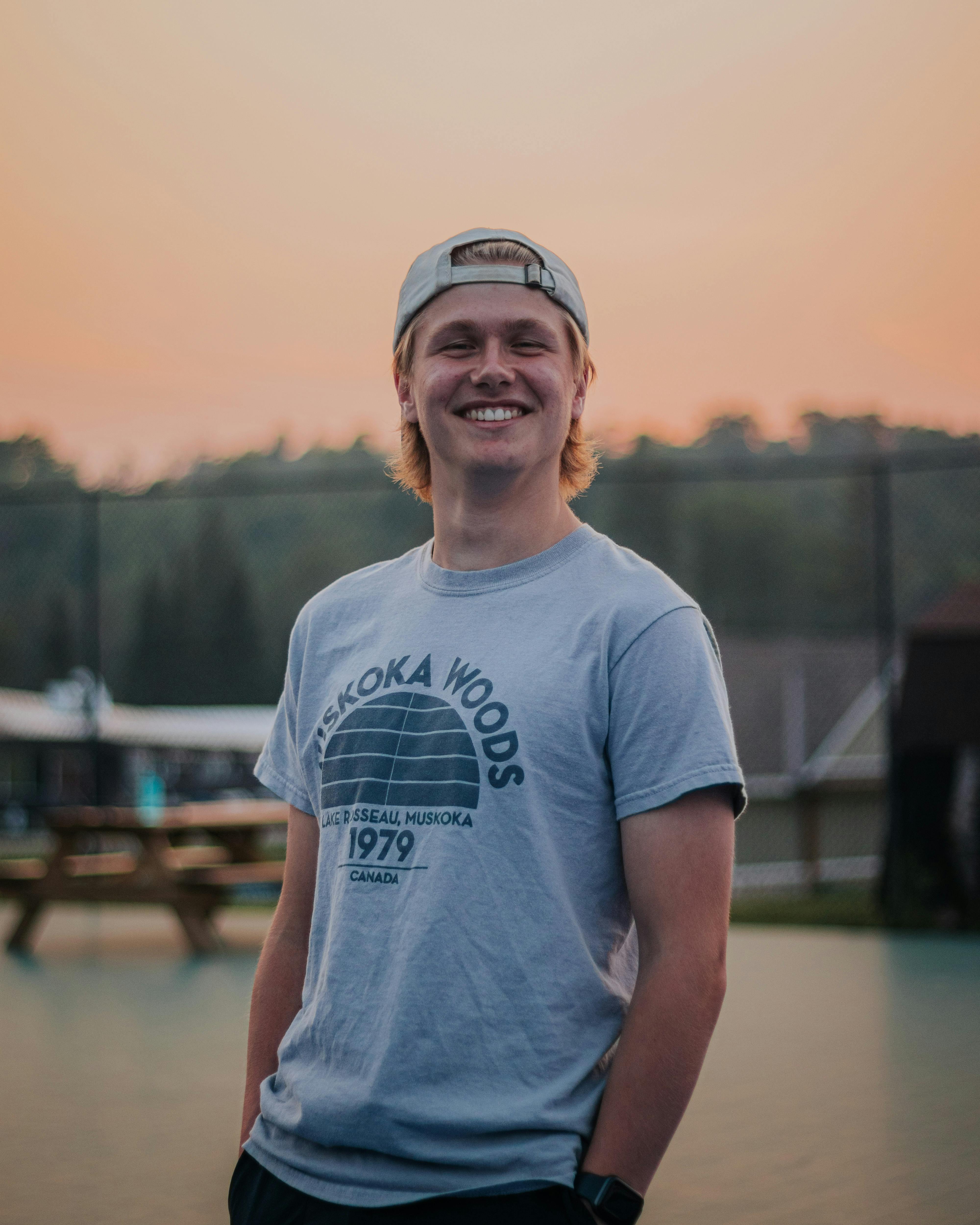A cheerful young man wearing a cap and t-shirt smiles outdoors during a sunset.