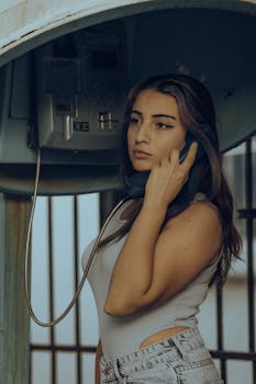 Young woman in a stylish outfit posing at a retro pay phone booth, exuding a vintage yet modern vibe.
