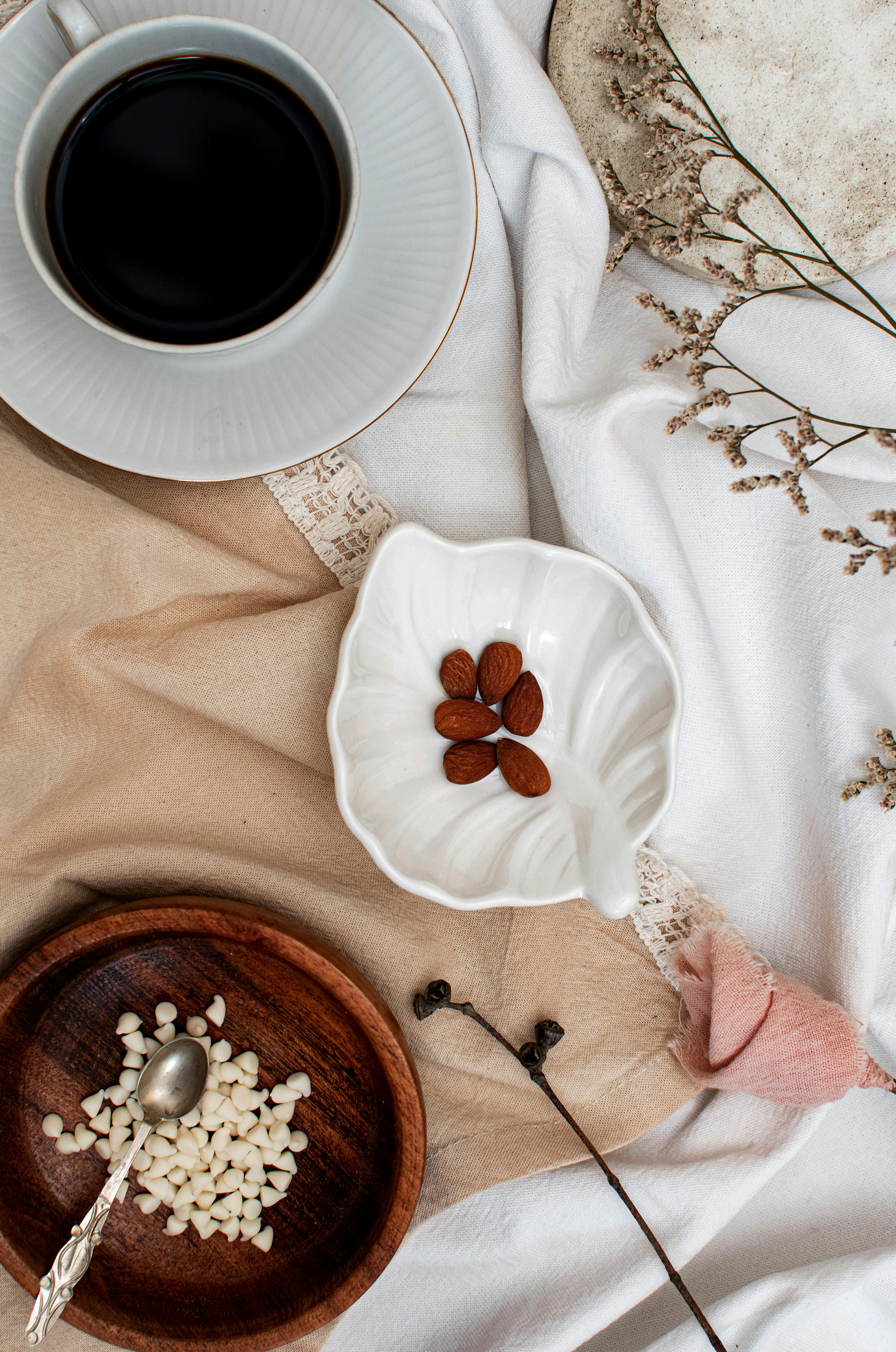 A cozy flat lay of coffee paired with almonds and white chocolate bits on elegant fabric.