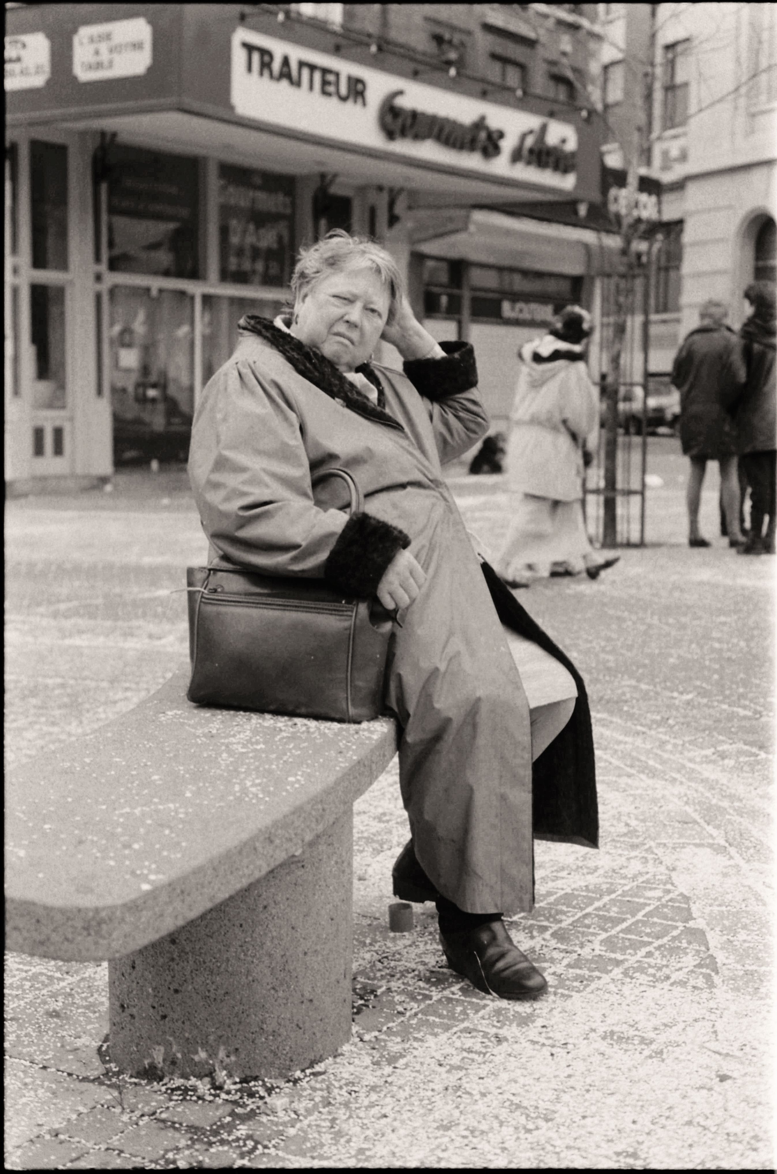 Black and white image of an elderly woman sitting on a bench, urban surroundings.
