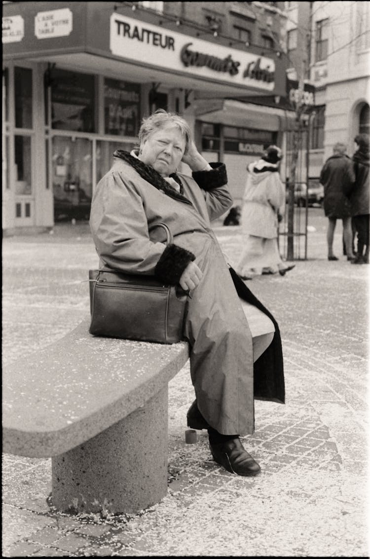 An Old Photograph Of An Elderly Woman Sitting On A Bench On The Street 