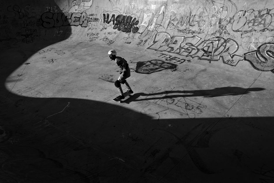 A skateboarder rides in a sunlit skatepark surrounded by urban graffiti, captured in black and white.