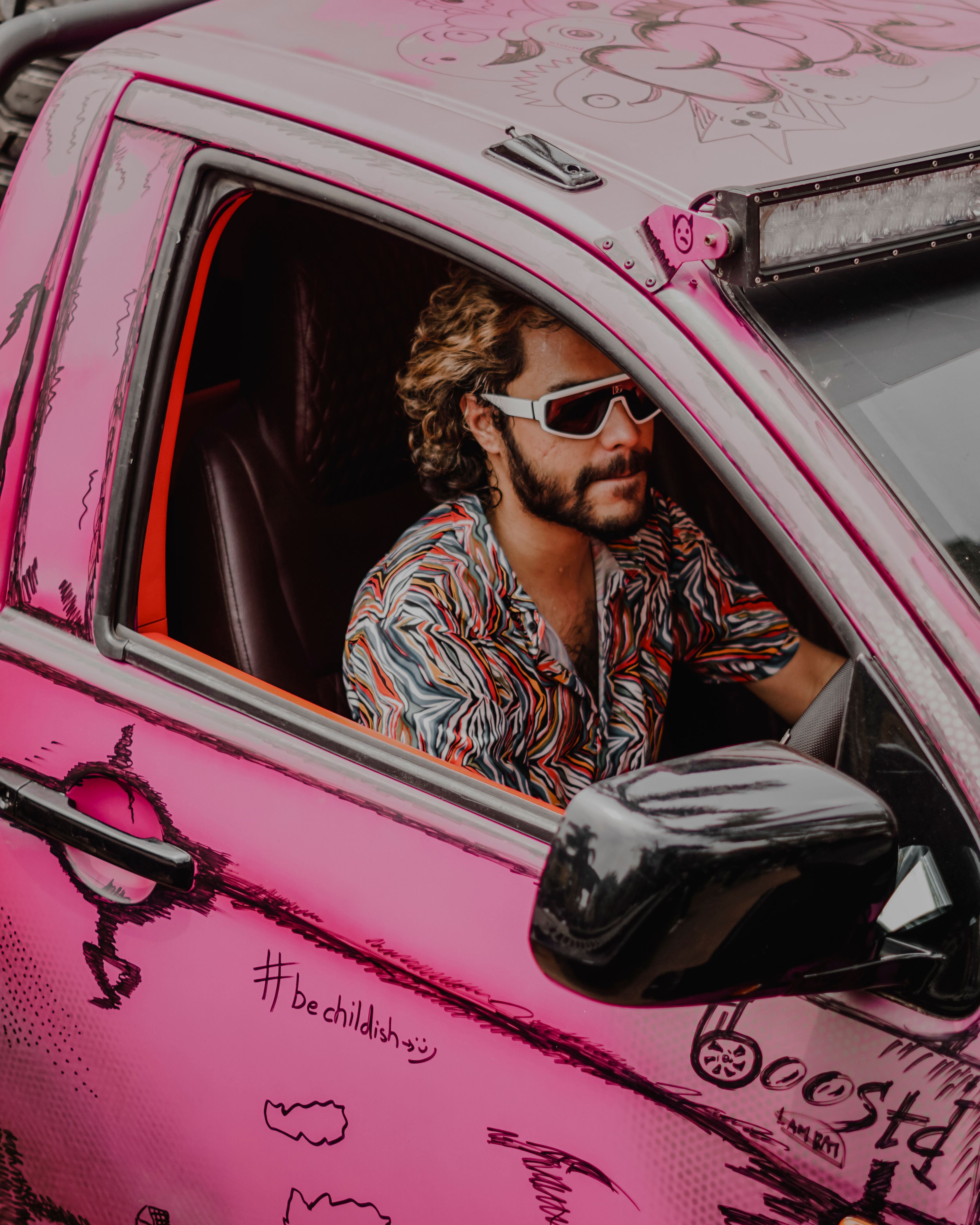 A young man with sunglasses and a beard drives a pink, graffiti-adorned truck.