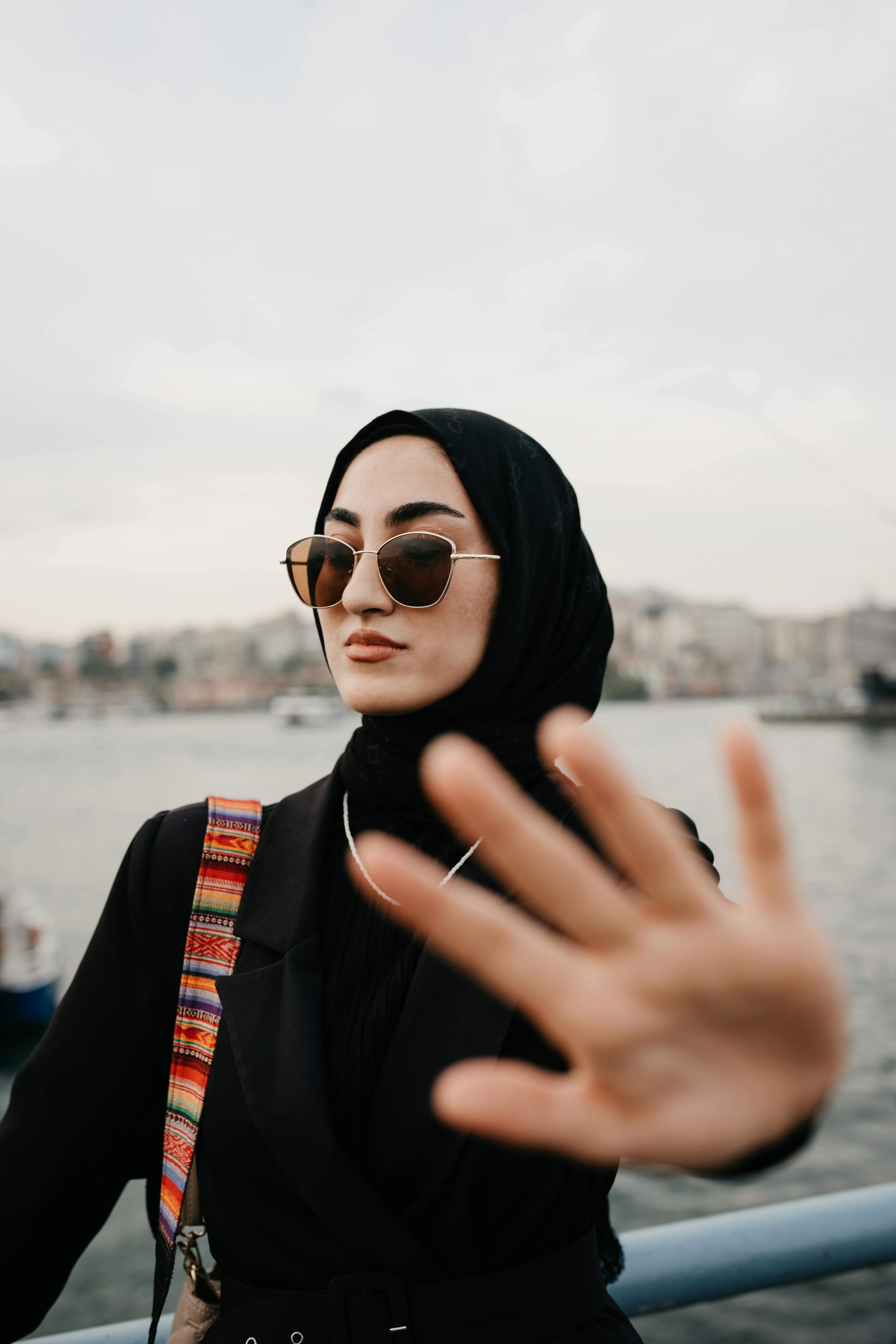 Fashion-forward woman in sunglasses and hijab standing by the waterfront, exuding confidence.