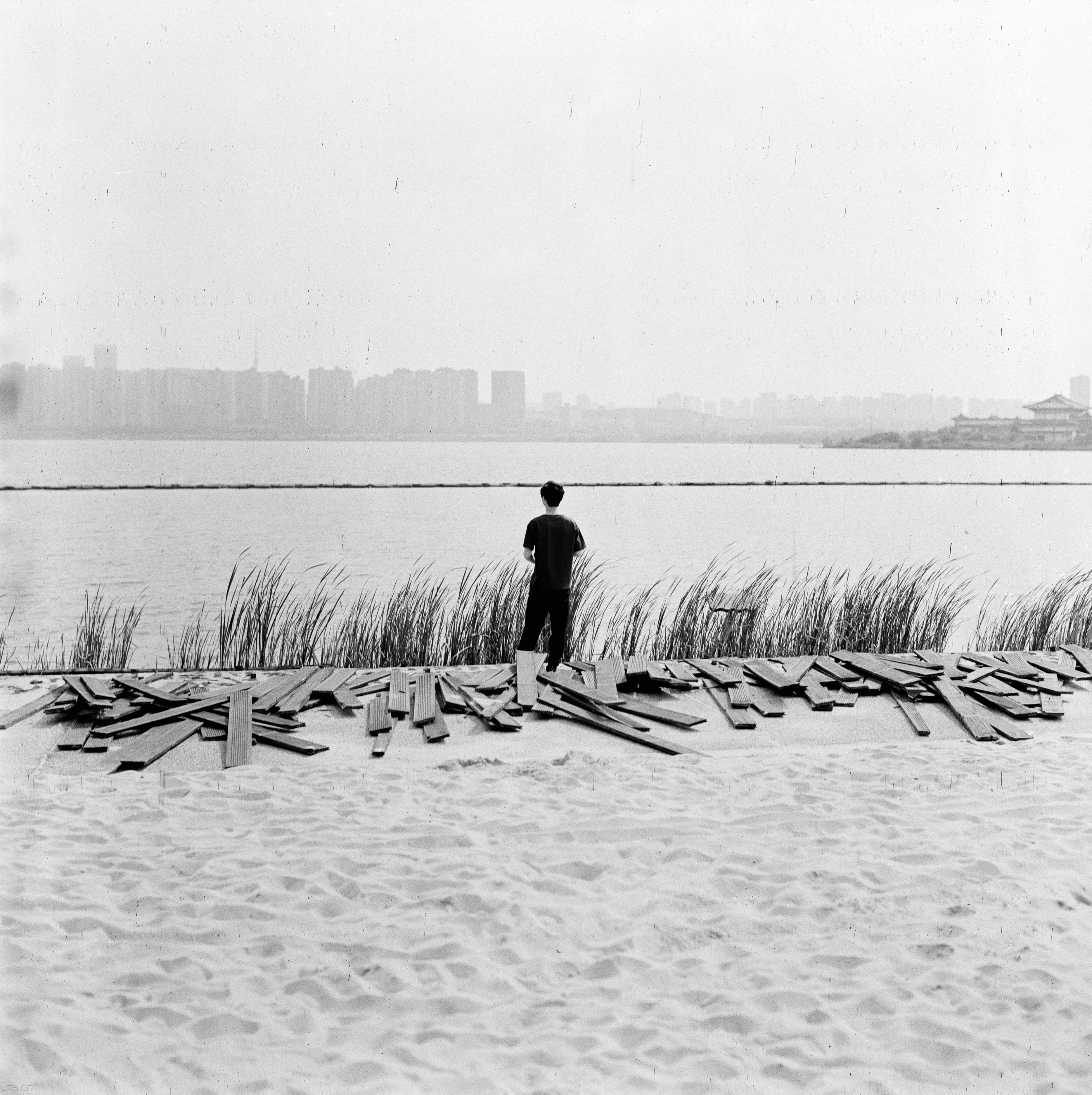 A lone man stands on a sandy beach, overlooking a serene waterfront and city skyline.