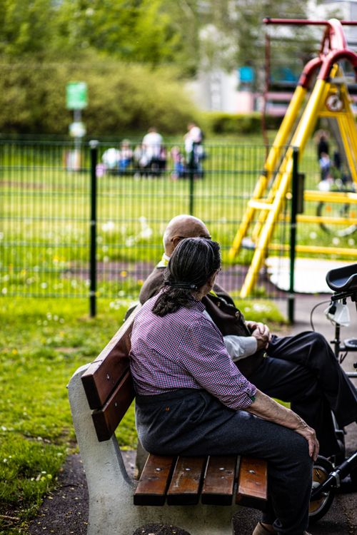 Free An elderly couple relaxes on a bench in a park, surrounded by green nature and playground equipment. Stock Photo