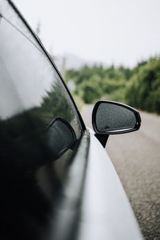 A car's side mirror covered in raindrops, reflecting a lush green landscape.