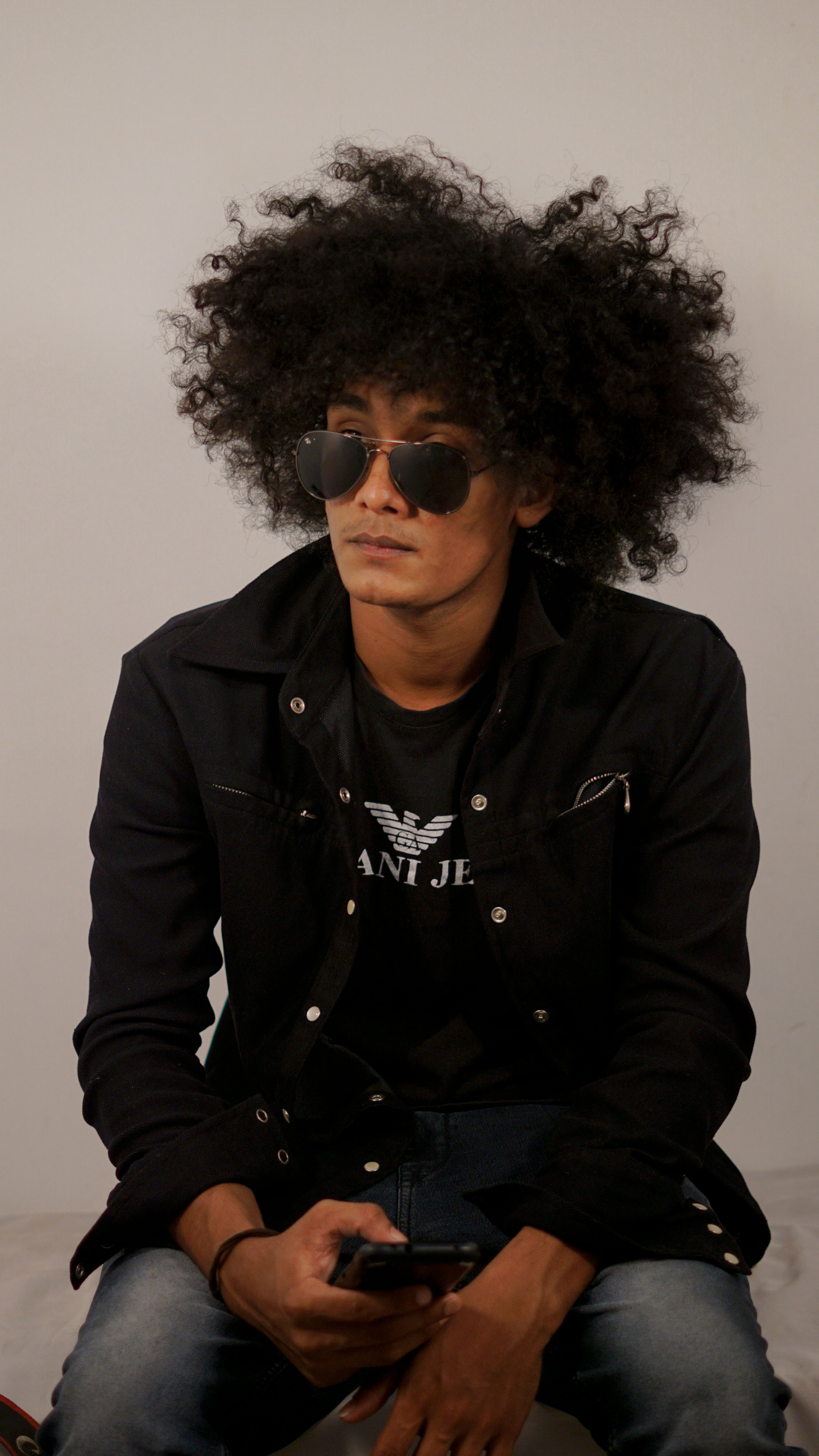 Young man with afro hairstyle wearing sunglasses and a black jacket, sitting casually indoors.
