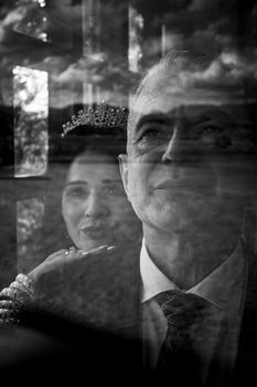 A reflective black and white portrait of a couple standing together, with a woman wearing a crown, seen through a window.