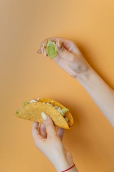 A close-up of hands holding a taco and lime on an orange background, emphasizing food freshness.