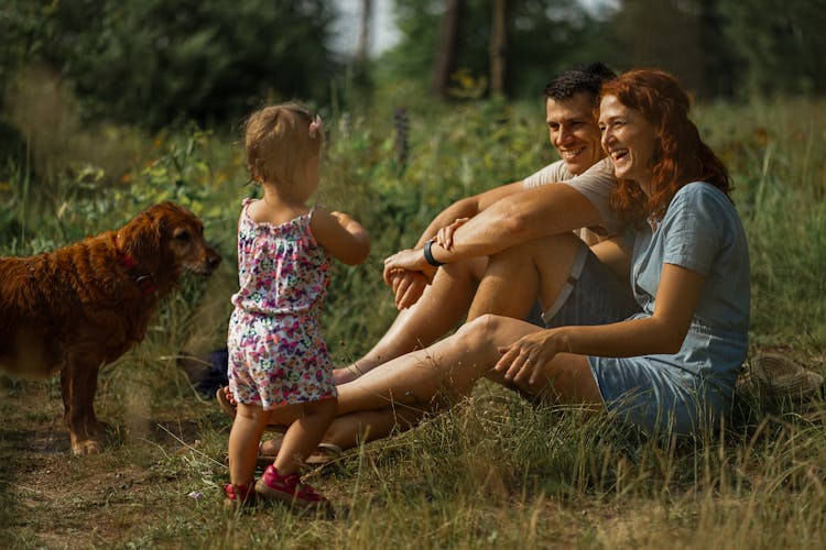 Happy Family With A Baby Daughter And Their Dog Relaxing On The Grass