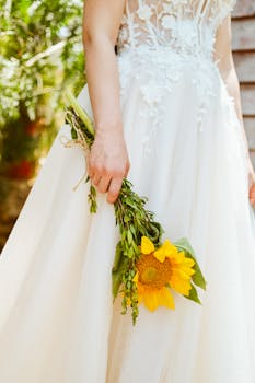 Elegant bride holding a sunflower bouquet, wearing a lace wedding dress in a sunlit outdoor garden.