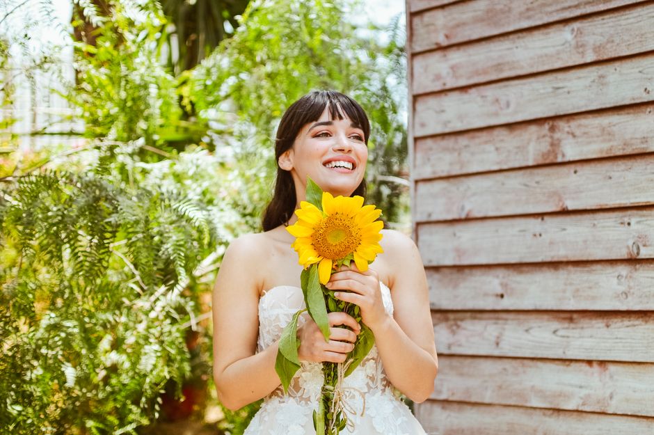 A joyful bride in a wedding dress holds a sunflower bouquet, standing against a rustic wooden wall outdoors.