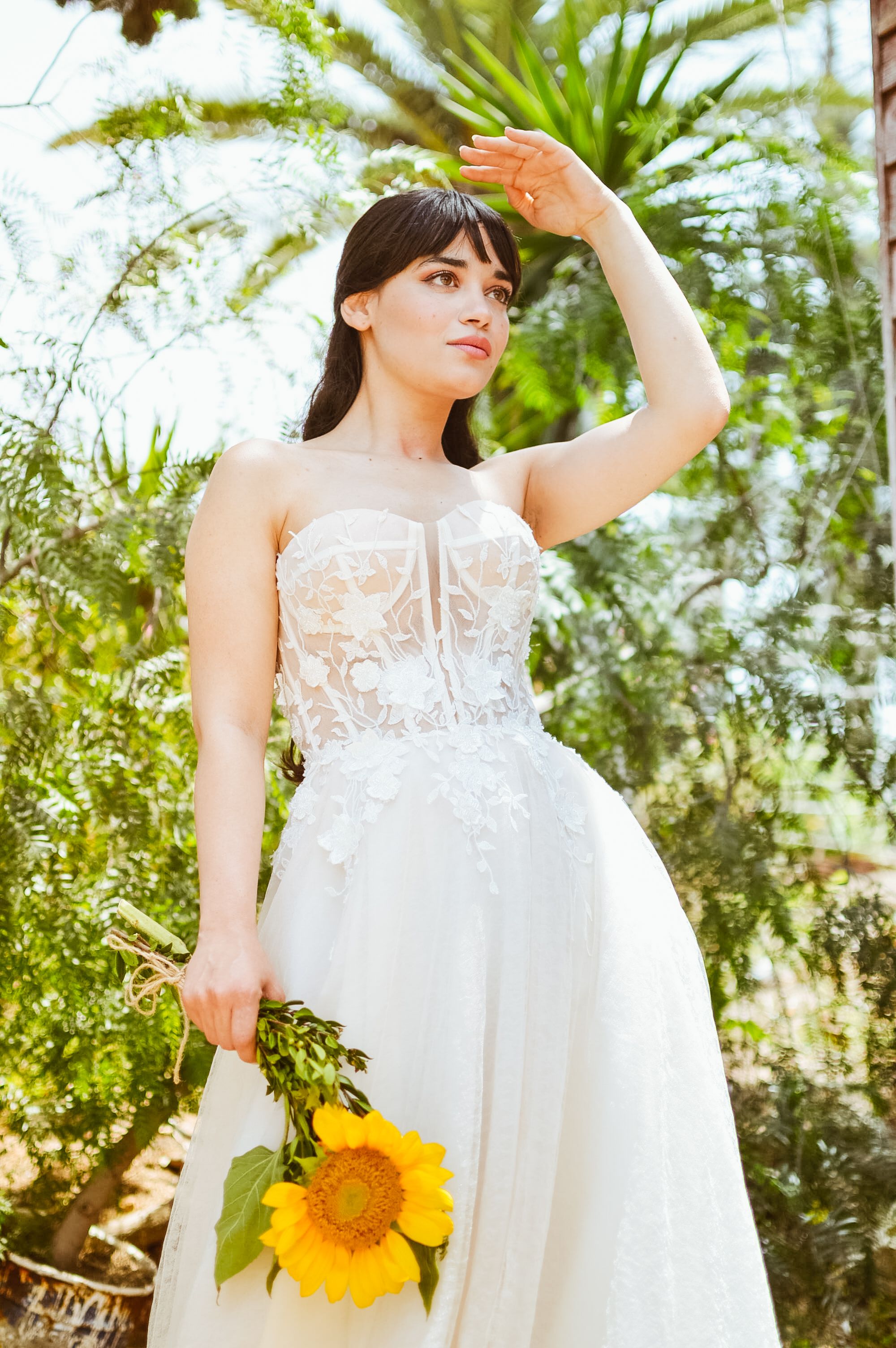 A stylish bride in a wedding dress holding a sunflower under bright sunlight.