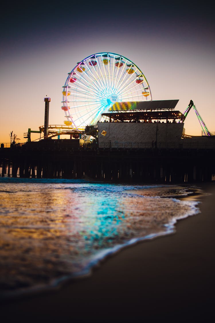 Illuminated Ferris Wheel On Sea Shore At Sunset