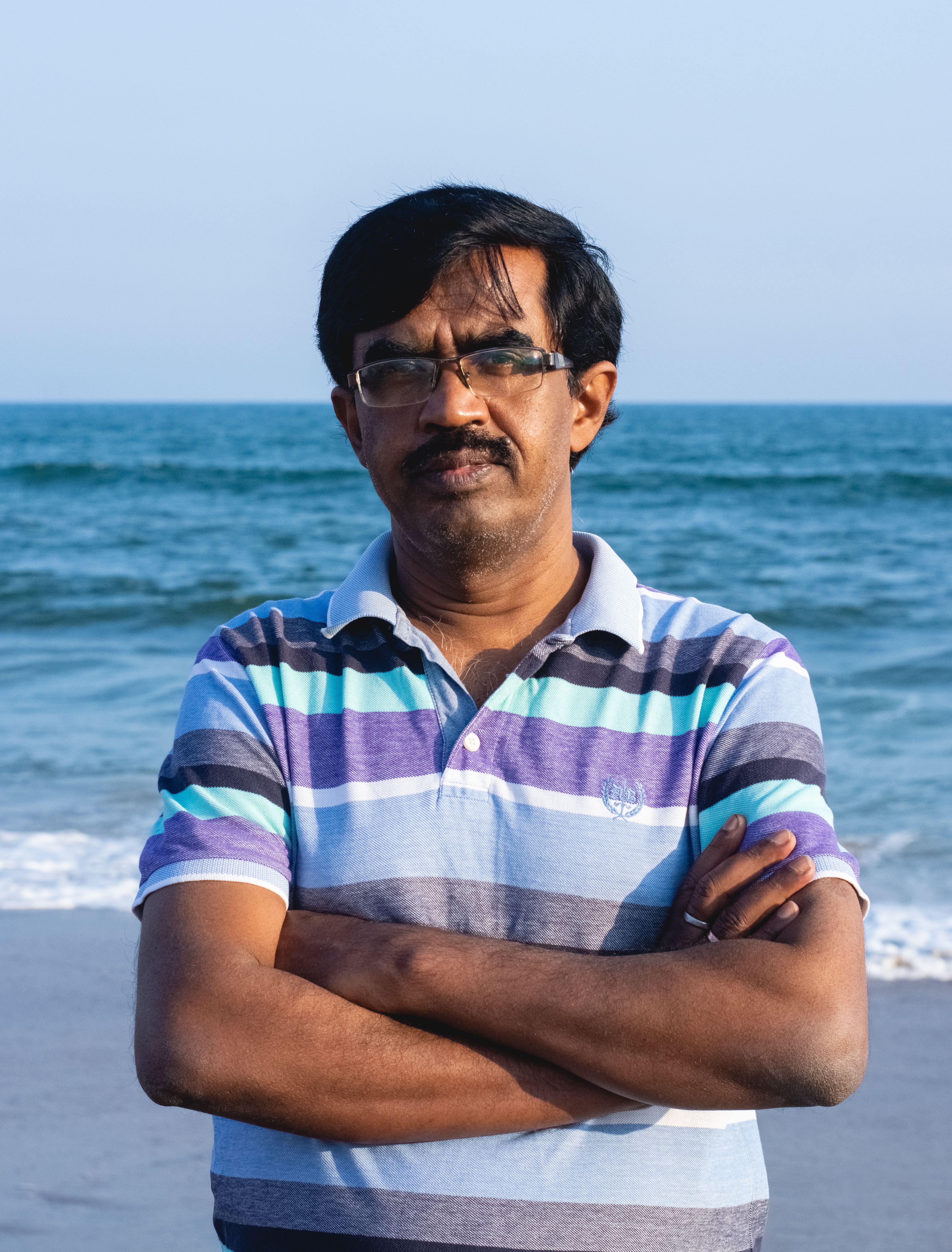 Portrait of a mature man standing at a beach with crossed arms and eyeglasses.