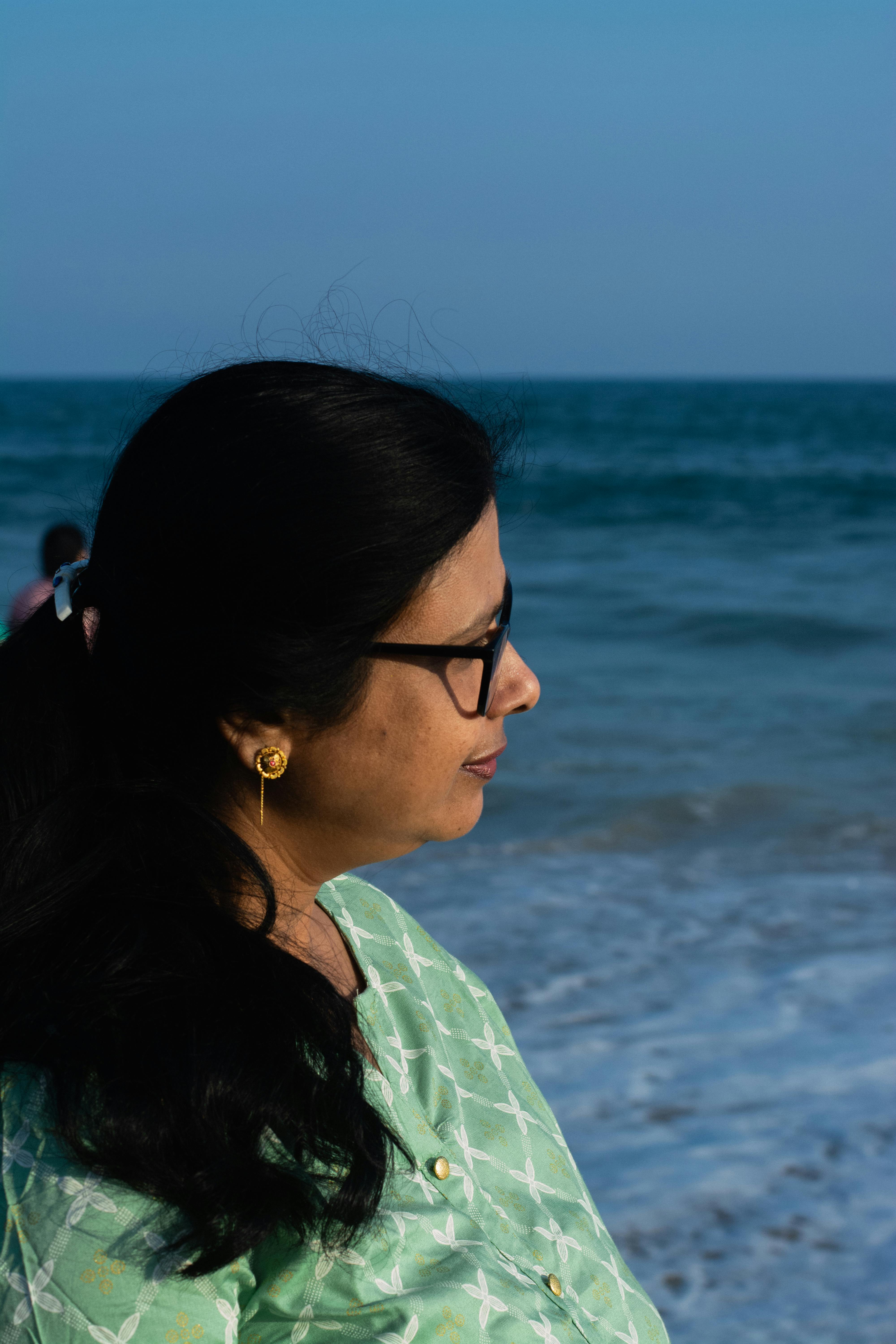 Profile of a woman with long black hair and eyeglasses standing by the sea shore on a bright day.