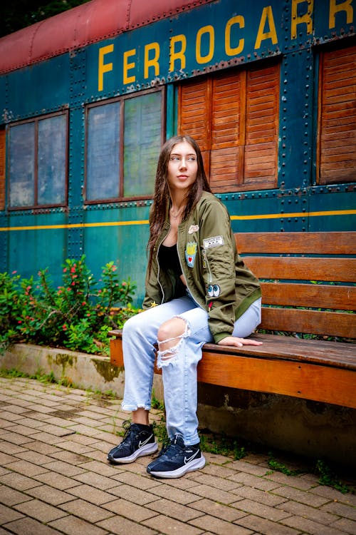 Young Woman in Ripped Jeans and Bomber Jacket Sitting on a Bench