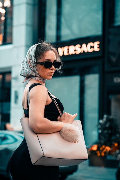 Free Elegant woman wearing a scarf and sunglasses, posing with a designer bag in a city setting. Stock Photo