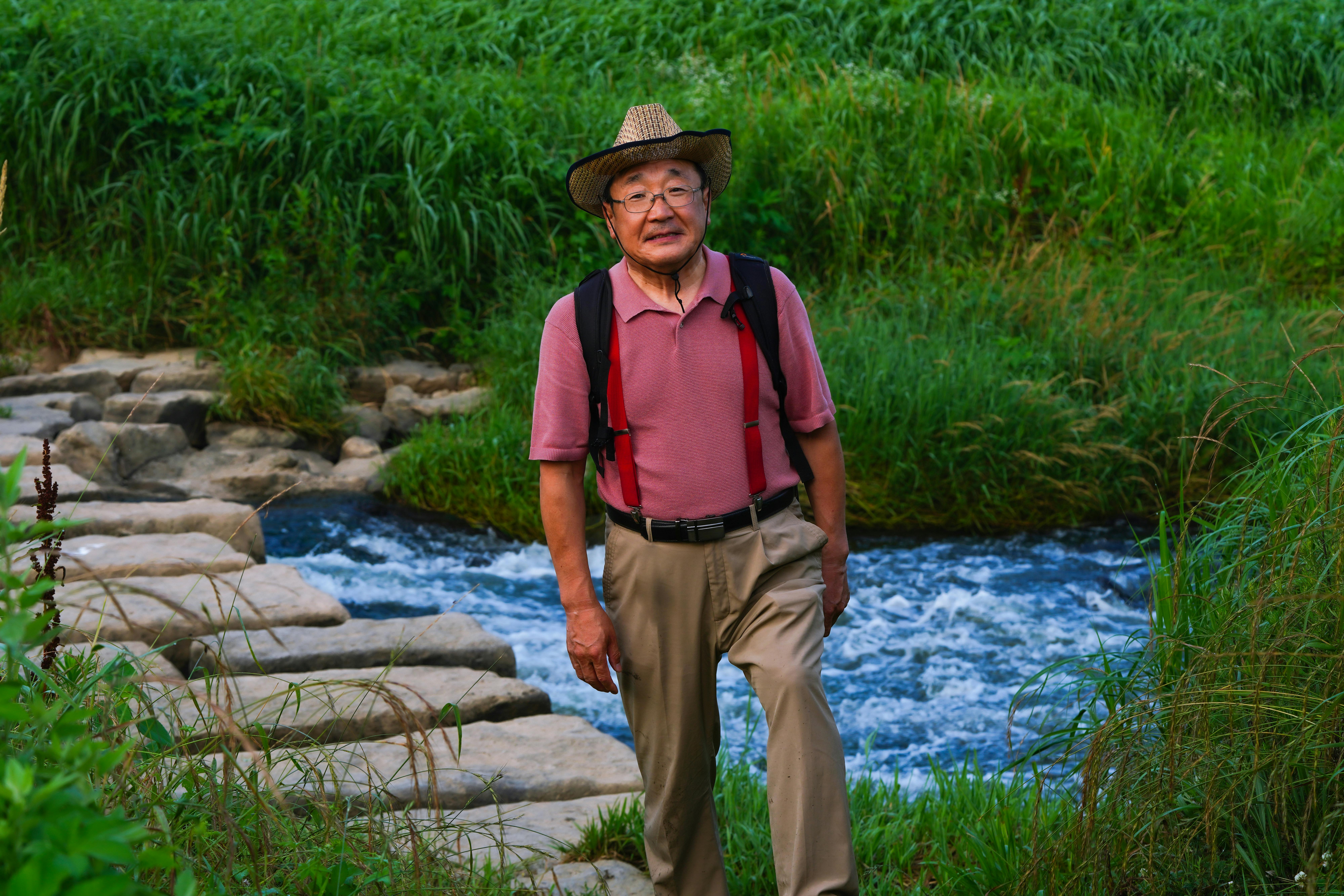 Elderly Asian man hiking on a stone path by a river, enjoying serene nature.