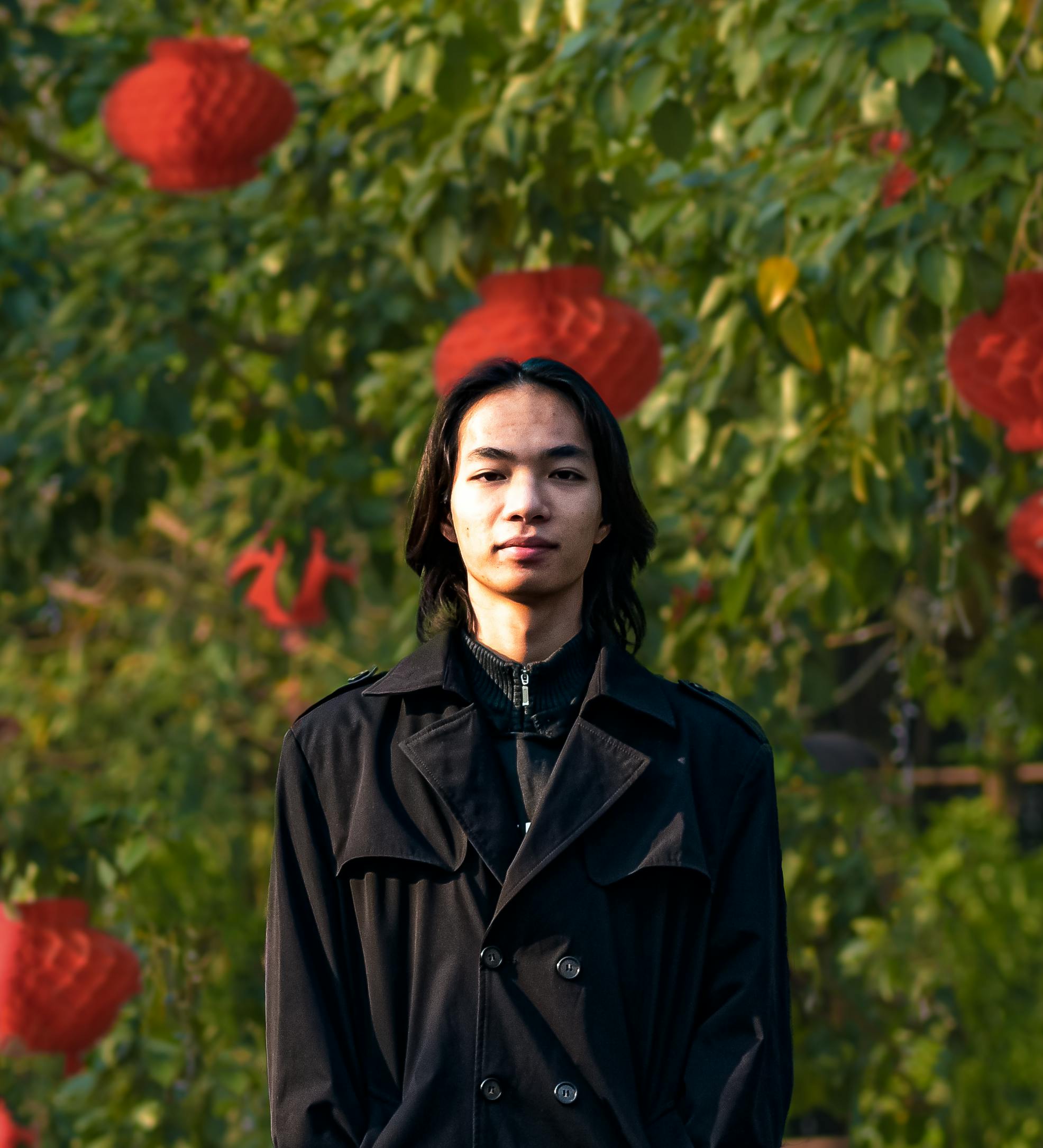 Man with long black hair in a stylish black coat stands outdoors with red lanterns in background.