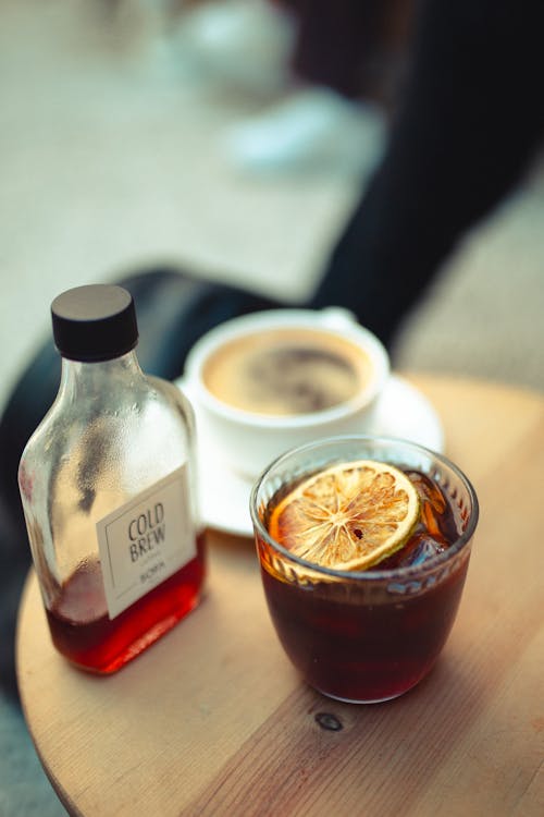 Drink with Ice Cubes and a Slice of Lemon Next to a Bottle and a Cup of Coffee