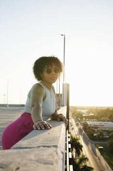 A woman in sunglasses leans on a rooftop railing enjoying the sunset view of the city.