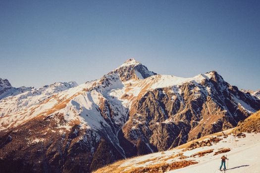 A lone skier traverses the snowy slopes of Dombay, Russia, beneath a clear blue sky.