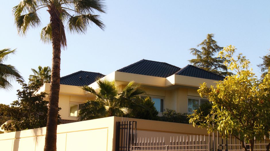 Contemporary Mediterranean villa with tiled roof and palm trees behind a perimeter wall under clear sky