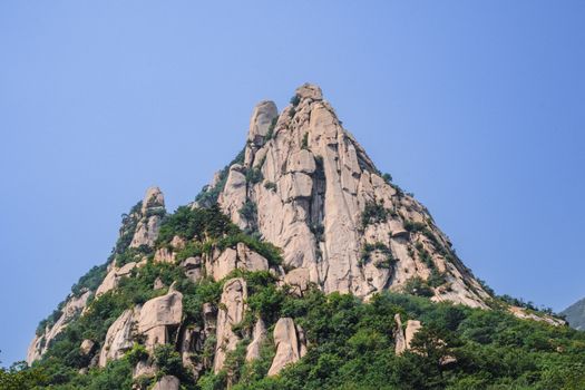 A stunning view of a rocky mountain peak under a clear blue sky, surrounded by lush greenery.
