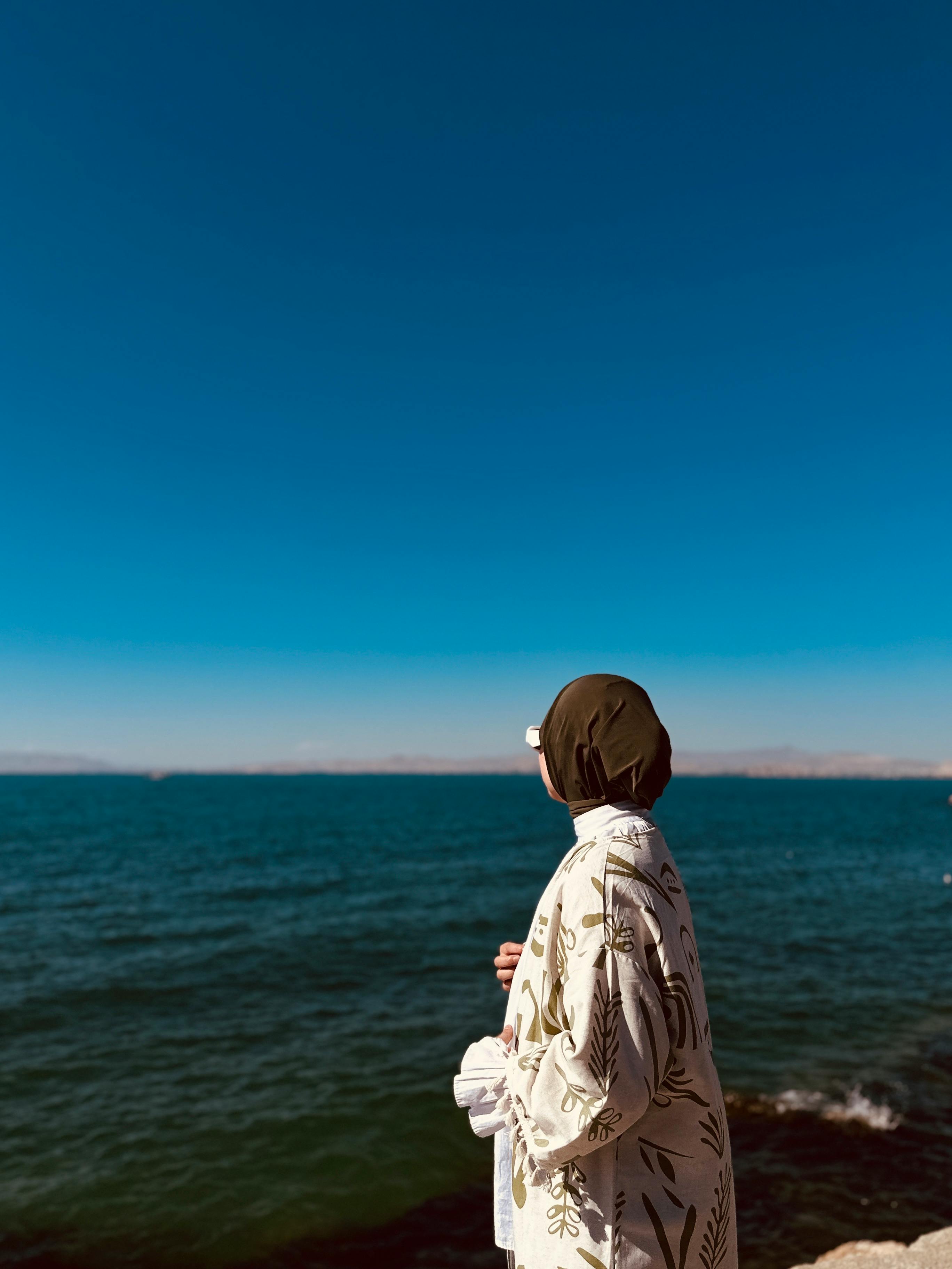 A woman in a hijab stands by the sea under a clear blue sky, embracing the serene coastal view.