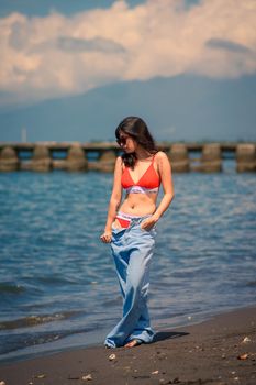 Young woman in red bikini and jeans enjoying a sunny day on the beach with ocean and mountain views.